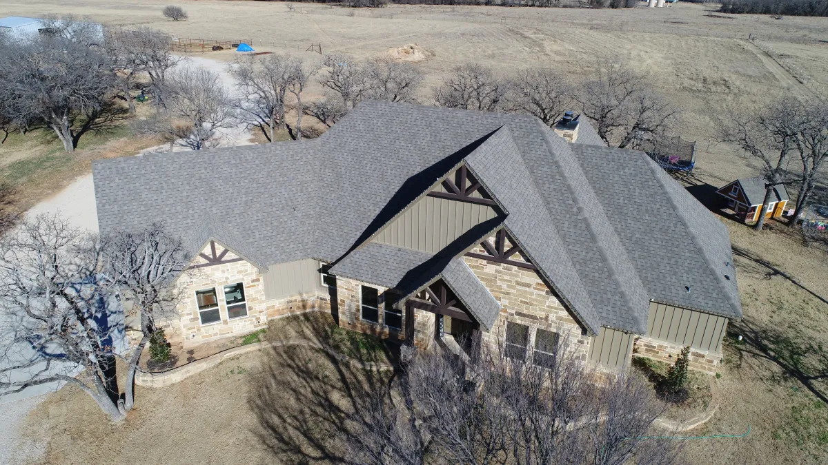 An aerial view of a large brown and beige stone house with a gray roof and several bare trees.